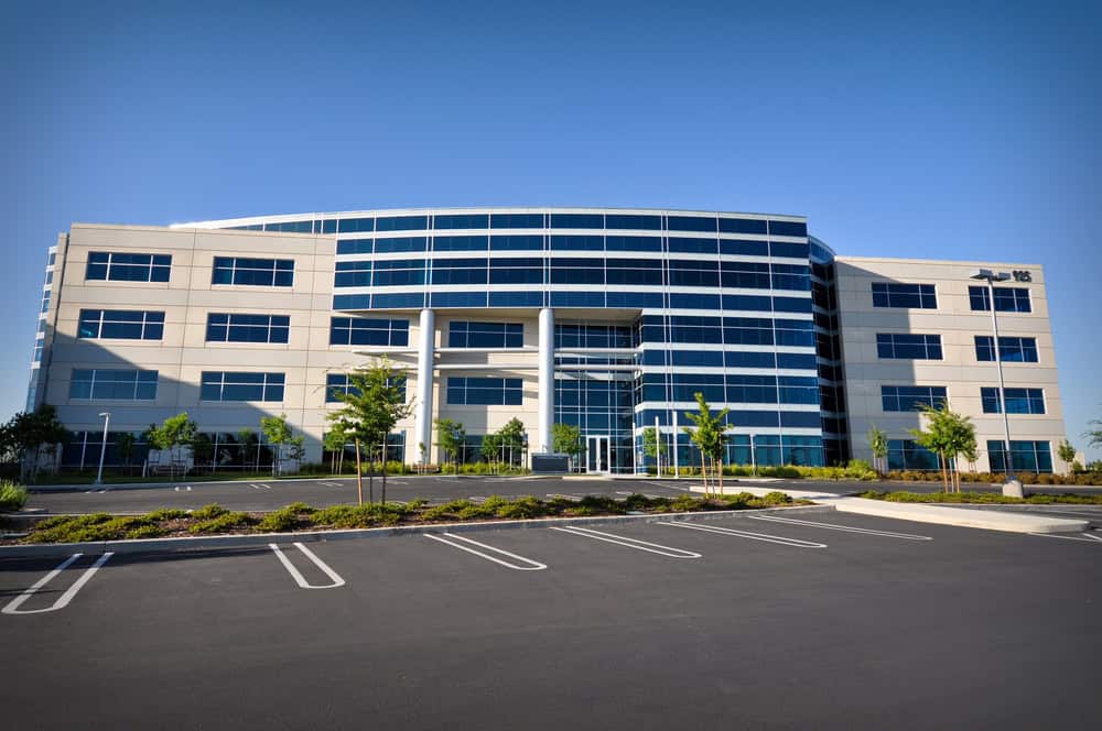 Modern four-story office building with glass windows, nestled among trees and an empty parking lot, under a clear blue sky. The adjacent landscaped area is meticulously maintained with top-notch lawn care, adding a refreshing touch of greenery to the sleek architectural design.