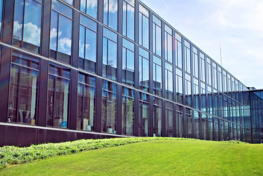 A glass-walled modern commercial building with reflective windows is bordered by a neatly maintained grassy lawn, thanks to expert fertilization services, on a sunny day.