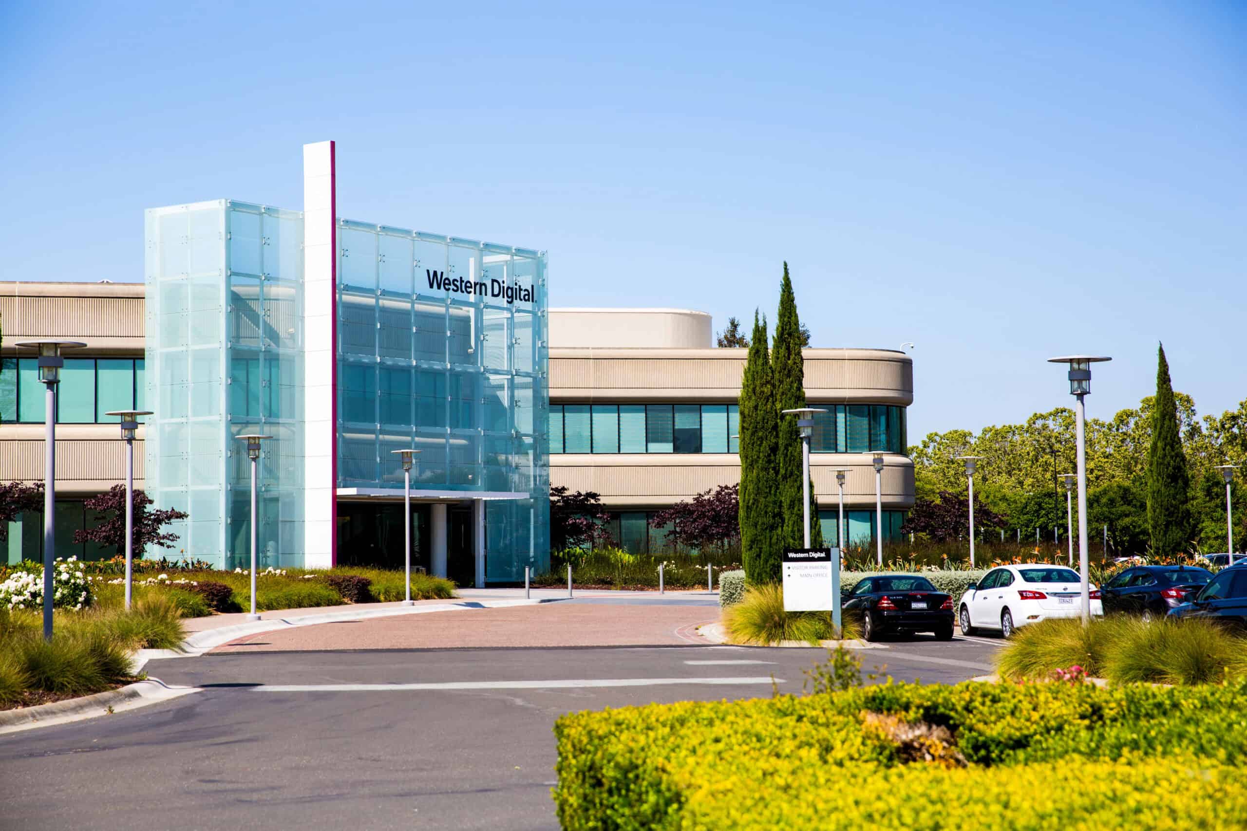 A modern office building with "Western Digital" on its glass facade, surrounded by well-maintained trees and a parking lot. The impeccable lawn care adds to the serene environment, making it an inspiring space for professionals.