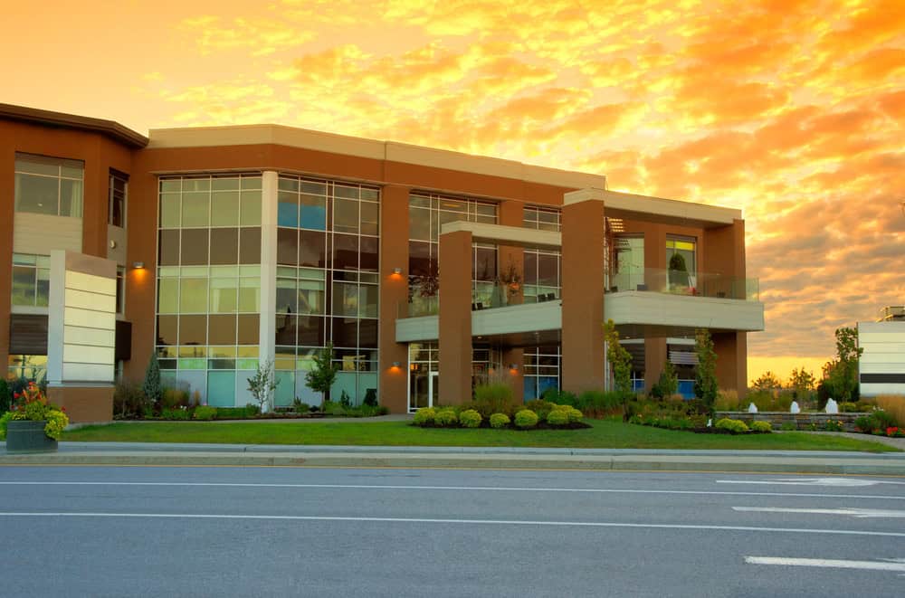 Modern medical facility with large glass windows and brown brick facade, set against a vibrant orange sunset sky, surrounded by landscaped greenery and a street in the foreground.