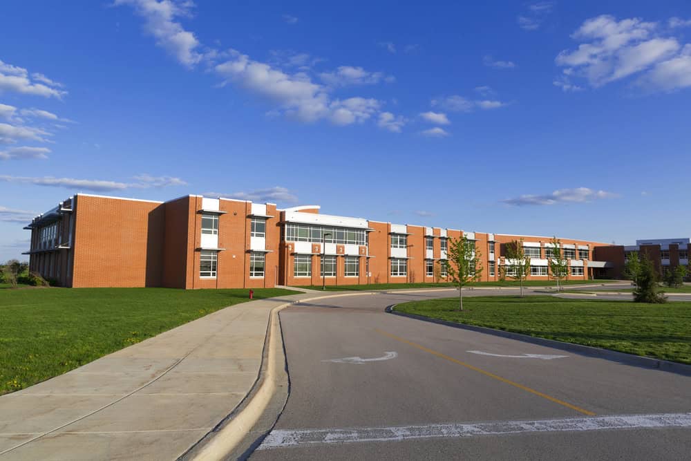 A large red-brick school building with numerous windows is set beside a curving road and a well-maintained grassy area, thanks to expert lawn care, all under a blue sky with scattered clouds.