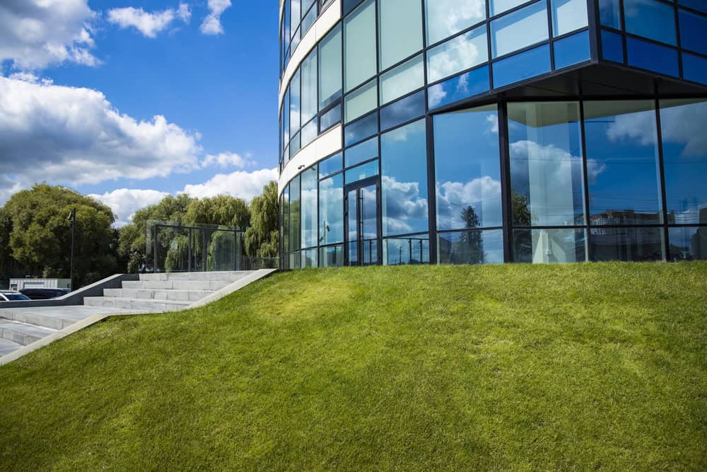 A modern glass commercial building with a curved facade reflects the sky and clouds, situated beside a green lawn under a partly cloudy blue sky.