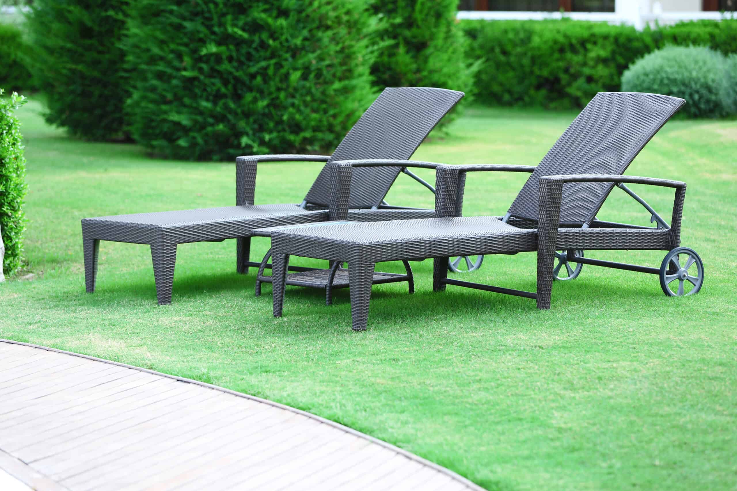 Two brown wicker lounge chairs and a matching table sit invitingly on a well-maintained grassy lawn near a wooden deck, with vibrant green bushes providing the perfect backdrop.