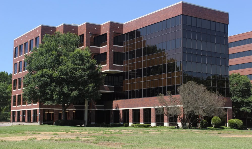 A four-story brick office building with large glass windows, nestled among trees and a grassy area designed for effective mosquito control, stands under a clear blue sky.