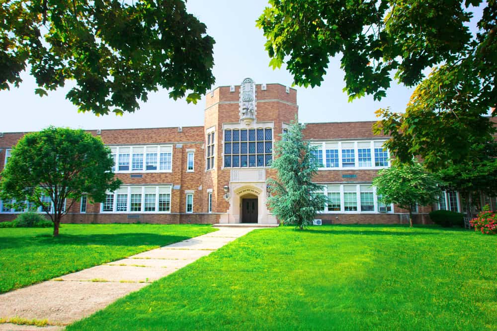 A brick school building with large windows and an arched entryway stands proudly, its central tower reaching for the sky. Surrounded by well-maintained lawns, thanks to meticulous lawn care, and vibrant trees under a clear blue sky.