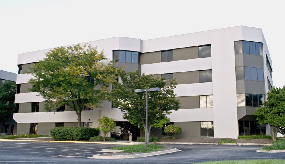 A four-story office building with a sleek white and gray exterior, featuring large windows, is surrounded by trees tended to with expert arborist services and a convenient parking lot.