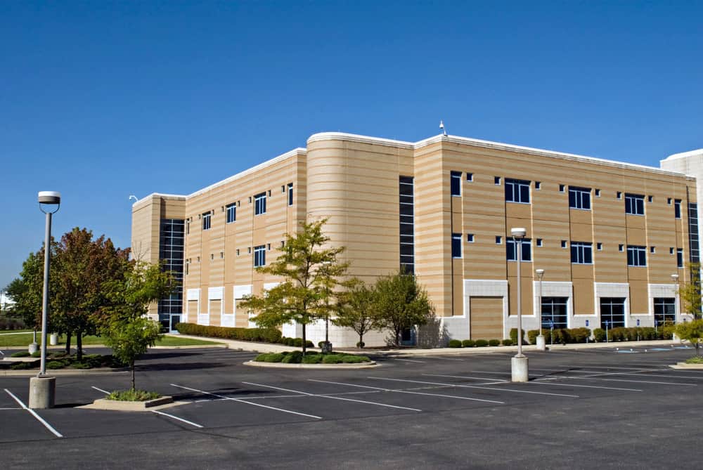 Three-story beige and white office building with large windows, possibly housing a medical facility, surrounded by an empty parking lot and small trees, under a clear blue sky.