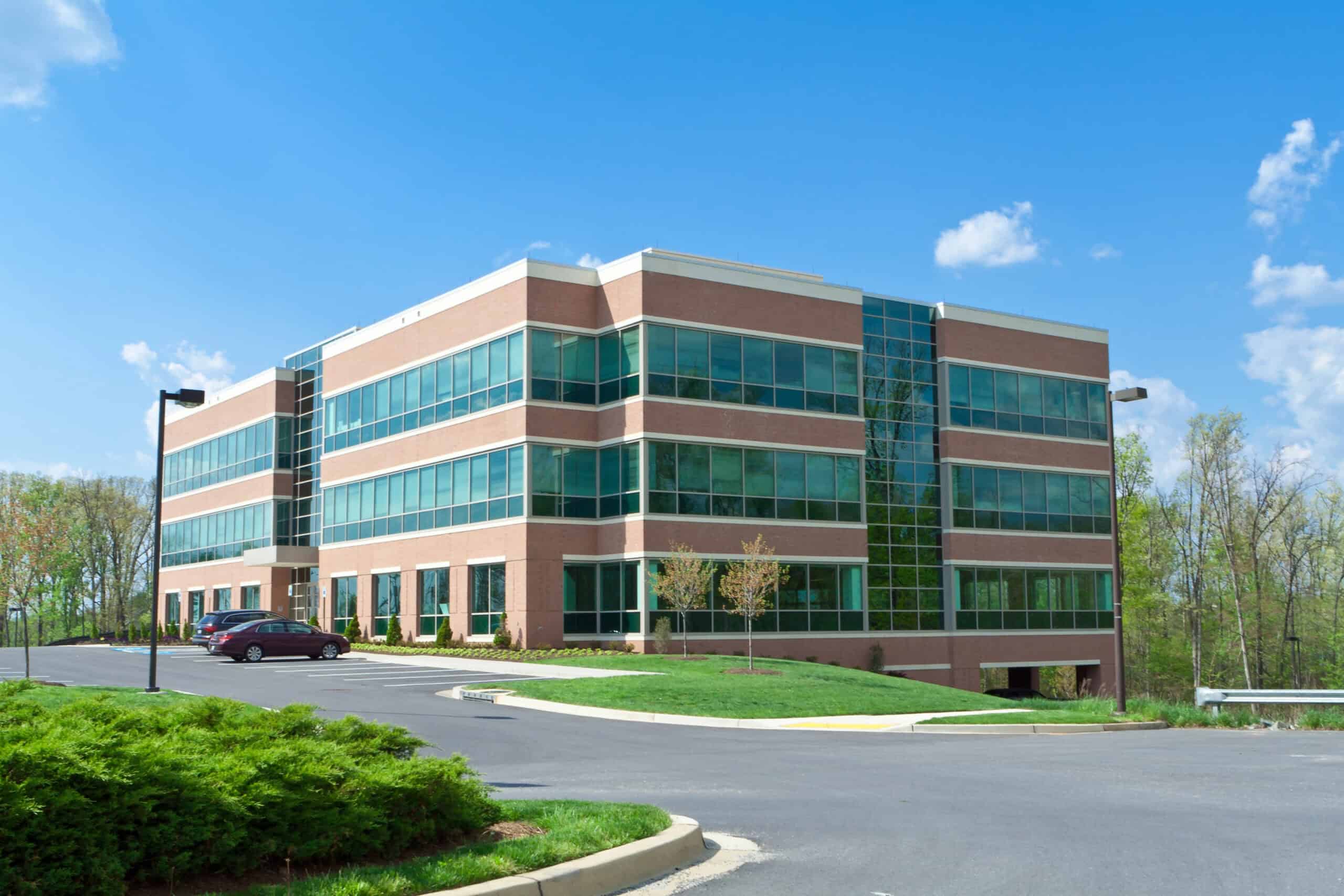 A modern, three-story office building with glass windows and a brick exterior rests against a clear blue sky. It's surrounded by trees, with a well-maintained lawn showcasing expert lawn care in the paved parking lot that has a few parked cars.