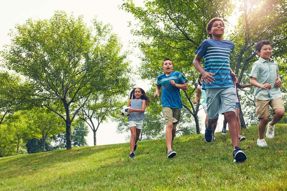Five children run and play with a soccer ball on a grassy hill in a park, enjoying the sunlight and trees while practicing summer safety for schools and camps.