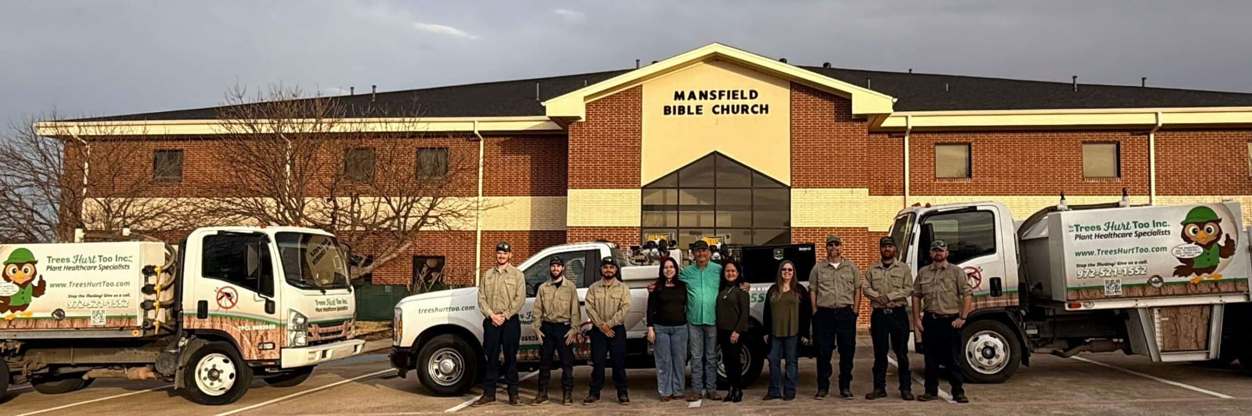 A group of people stands in front of three trucks displaying TX | Trees Hurt Too and Pest Control Tarrant County, Inc. logos, with the Mansfield Bible Church building in the background.