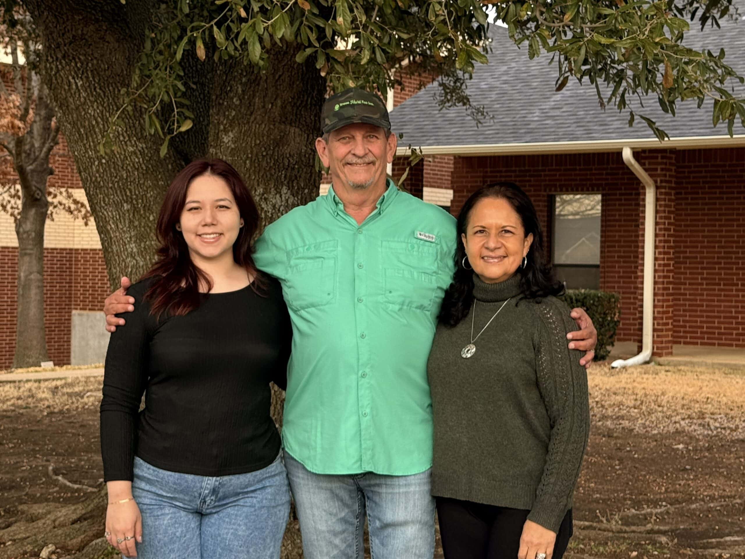 Three people stand together outside in front of a tree and a brick building, smiling at the camera-capturing the friendly spirit of Trees Hurt Too, Inc. Learn more on our About Us page.